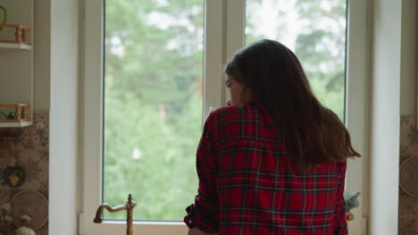 upset woman stands by window in kitchen. dark haired lady suffers from seasonal depression disorder staying alone at home. woman with mental health problems