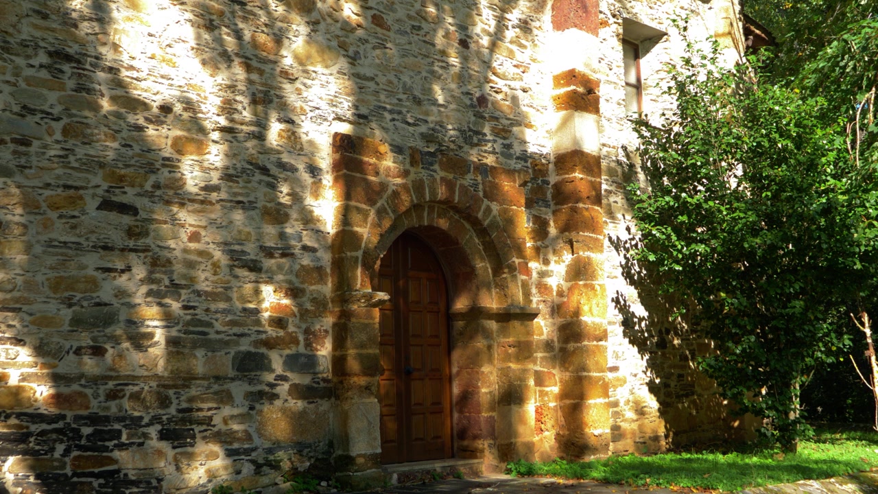 Vista esterna ravvicinata dell'ingresso ad arco in pietra di San Miguel de Xagoaza, incorniciato da alberi e muratura invecchiata, nel complesso storico della chiesa rurale di Valdeorras, Galizia