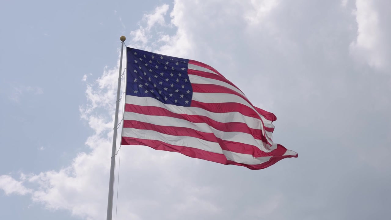 Et stort amerikansk flag på en flagstang, baggrundsbelyst af solen, blafrer i vinden på en delvist skyet sommerdag i slow motion.