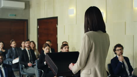 rear view of businesswoman speaking on the podium in a conference room and demonstrating video presentation on the screen behind her