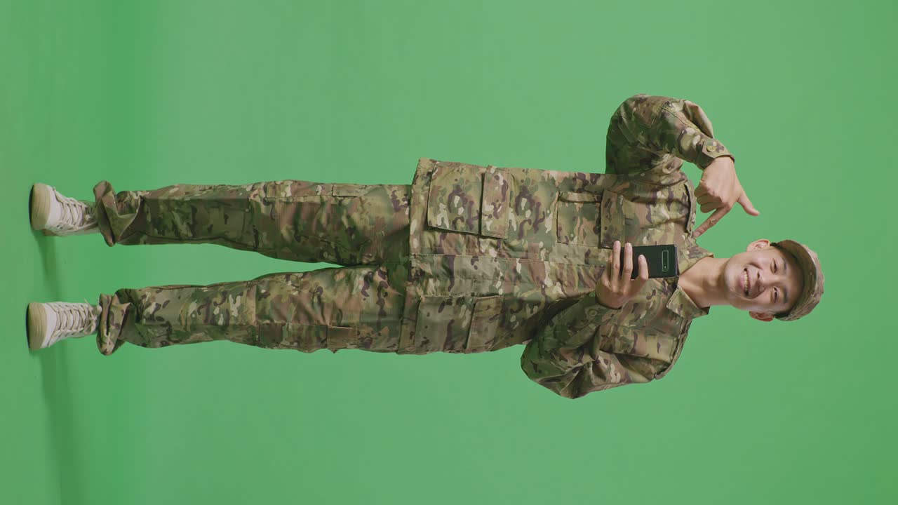 Full Body Of Asian Man Soldier Smiling And Pointing To Smartphone In His Hand While Standing In The Green Screen Background Studio