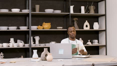 american clerk sitting at desktop in front of laptop counting banknotes in a pottery shop