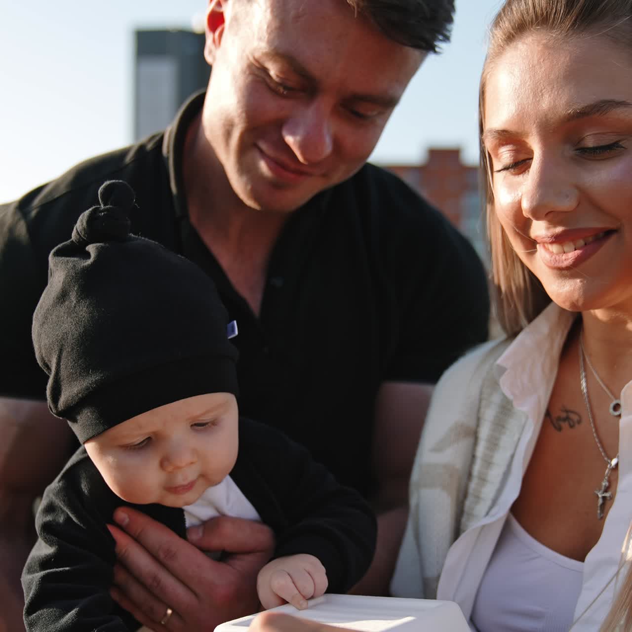 Caucasian family standing together outdoors. Man holding an infant boy interested in the box opened by mom. Close up. Vertical view