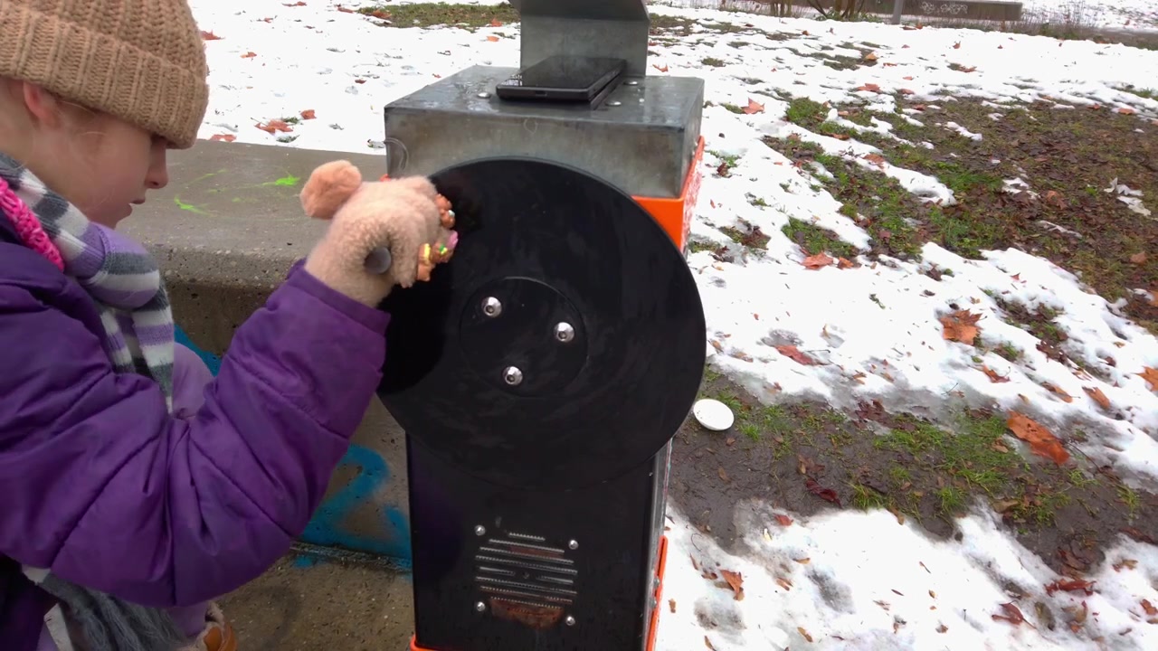Side view tracking shot of a young child turning a kinetic hand crank wheel at an interactive park science game display. Snow covers the playground lawn at Varosliget city park in Budapest Hungary.