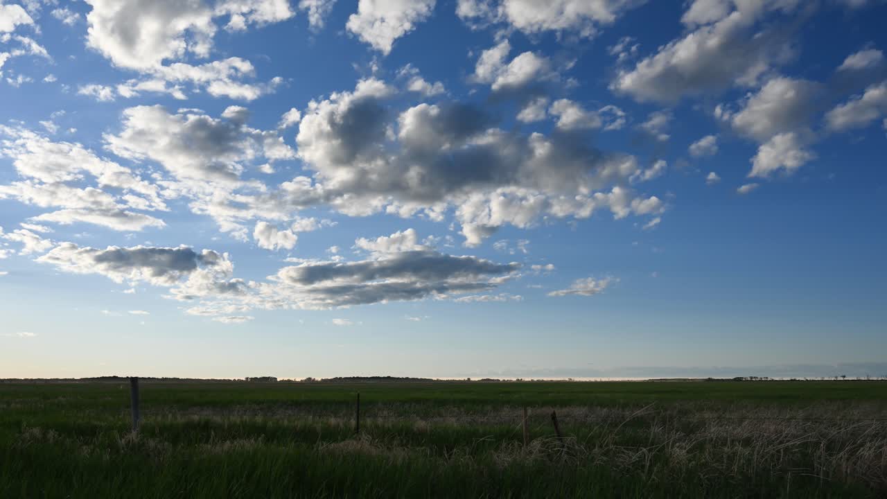 Time lapse of multiple layers of white blowing clouds moving across a bright blue sky above a farm field with wooden fence posts.
