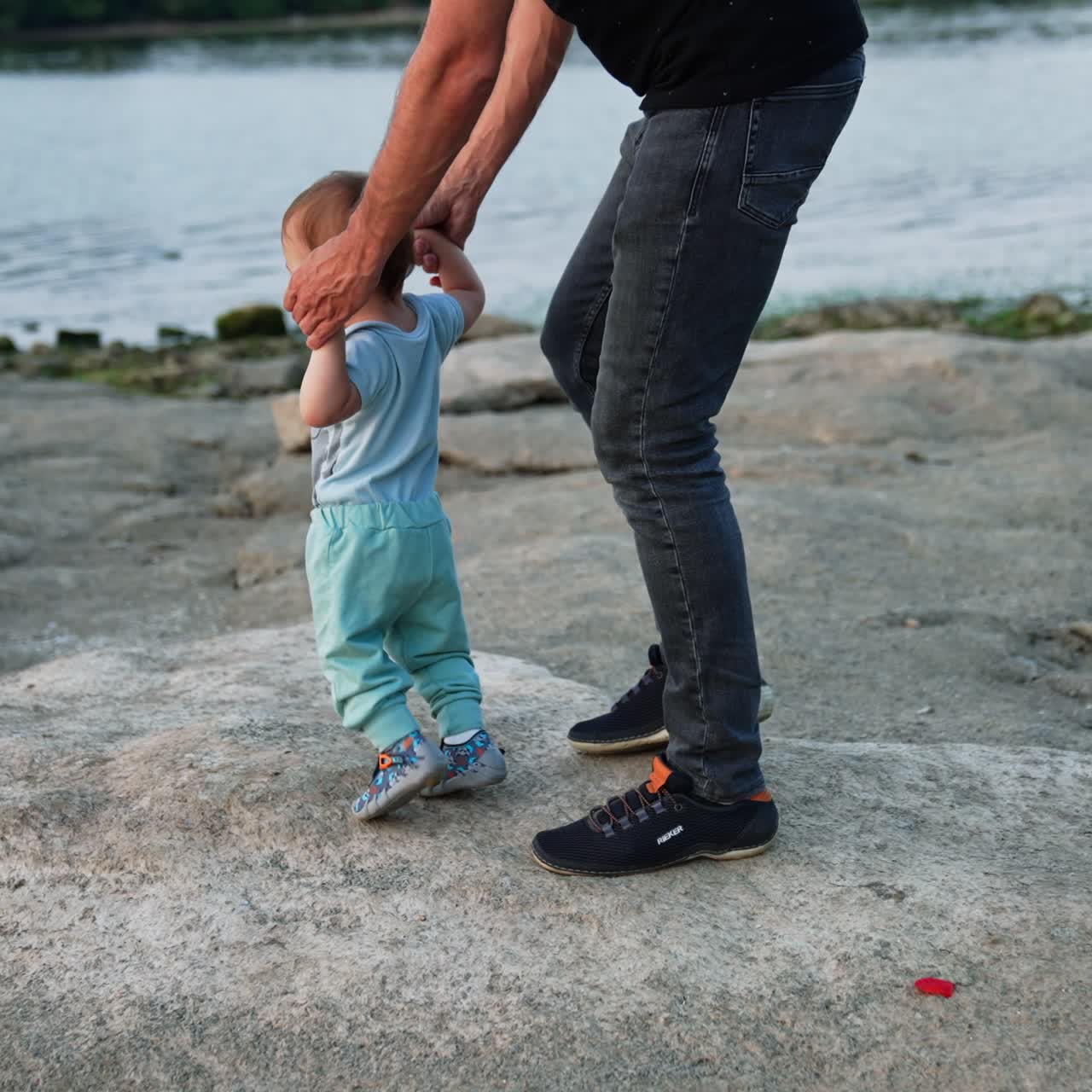 Uomo con jeans che accompagna il suo figlioletto che impara a camminare. Bellissimo neonato con vestiti blu che muove i primi passi sulle pietre. Sponda del fiume sullo sfondo