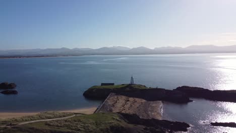 Vista-Aérea-De-La-Idílica-Isla-De-Ynys-Llanddwyn-Con-La-Brumosa-Cordillera-De-Snowdonia-A-Través-Del-Brillante-Mar-Irlandés-Hacia-El-Faro