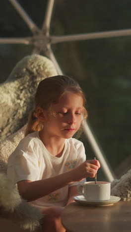 relaxed child stirs drink with spoon at glamping. quiet girl has leisurely breakfast sitting at table in transparent tent during holiday slow motion