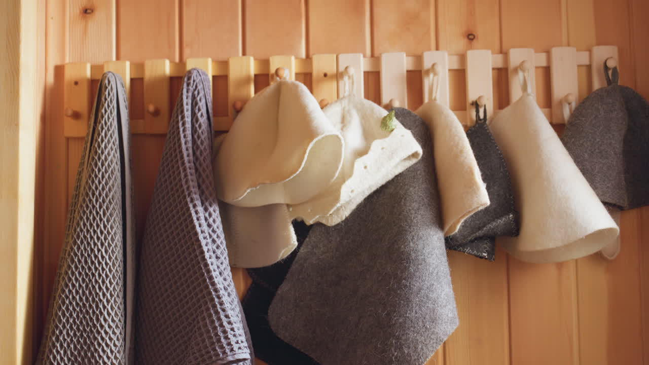 wooden sauna corner featuring hook rail with hanging waffle towels and felt hats, illuminated by warm daylight through nearby window, highlighting textured pine panels