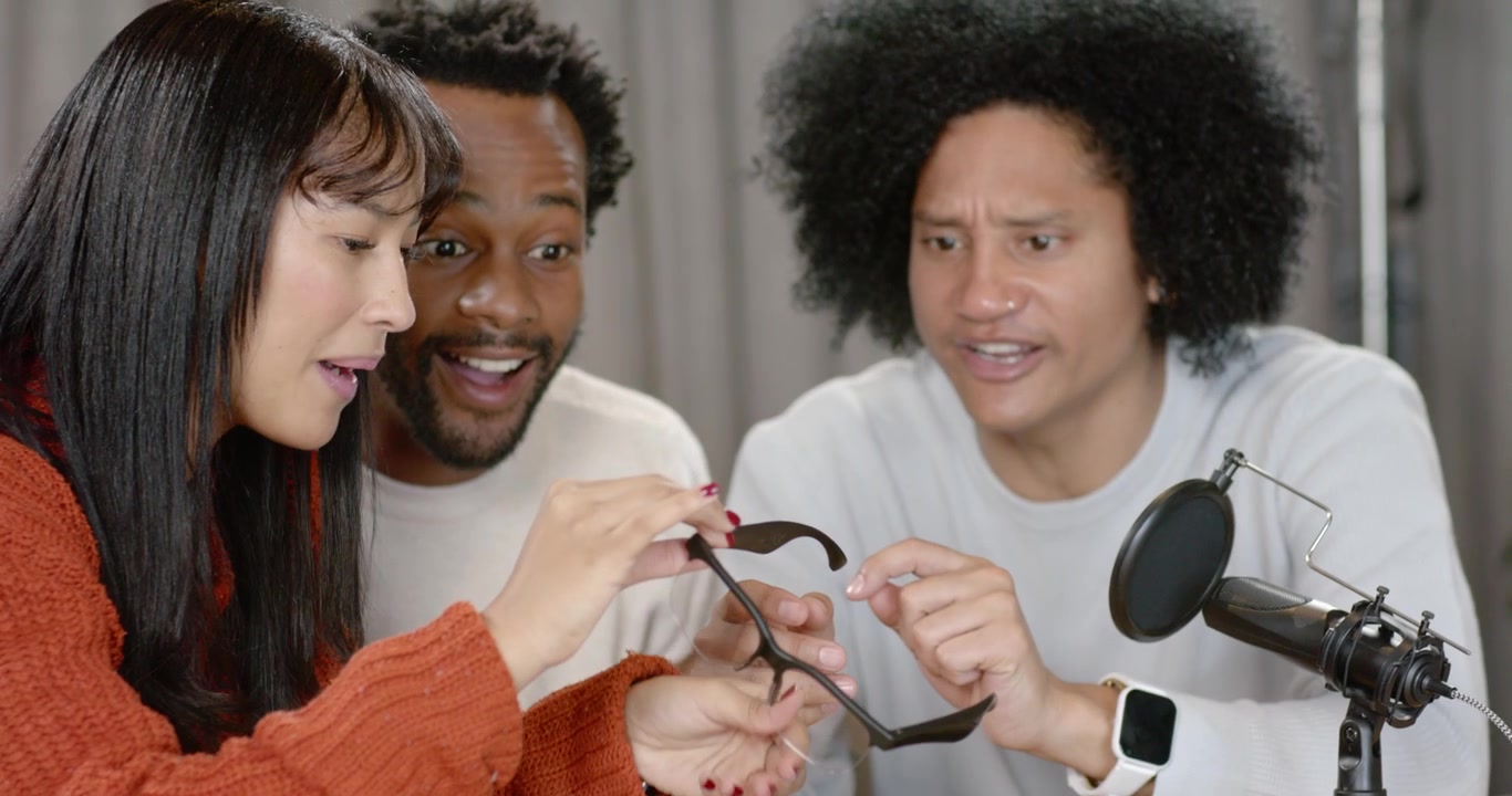 Diverse hosts leaning after Asian woman showing black smart glasses on table for review with mic