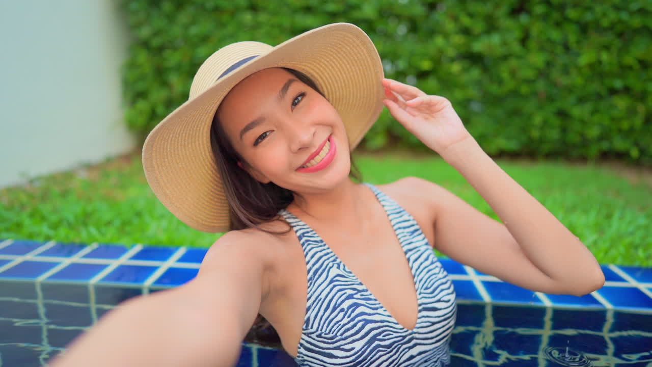 A young Asian woman in a resort swimming pool poses for a selfie