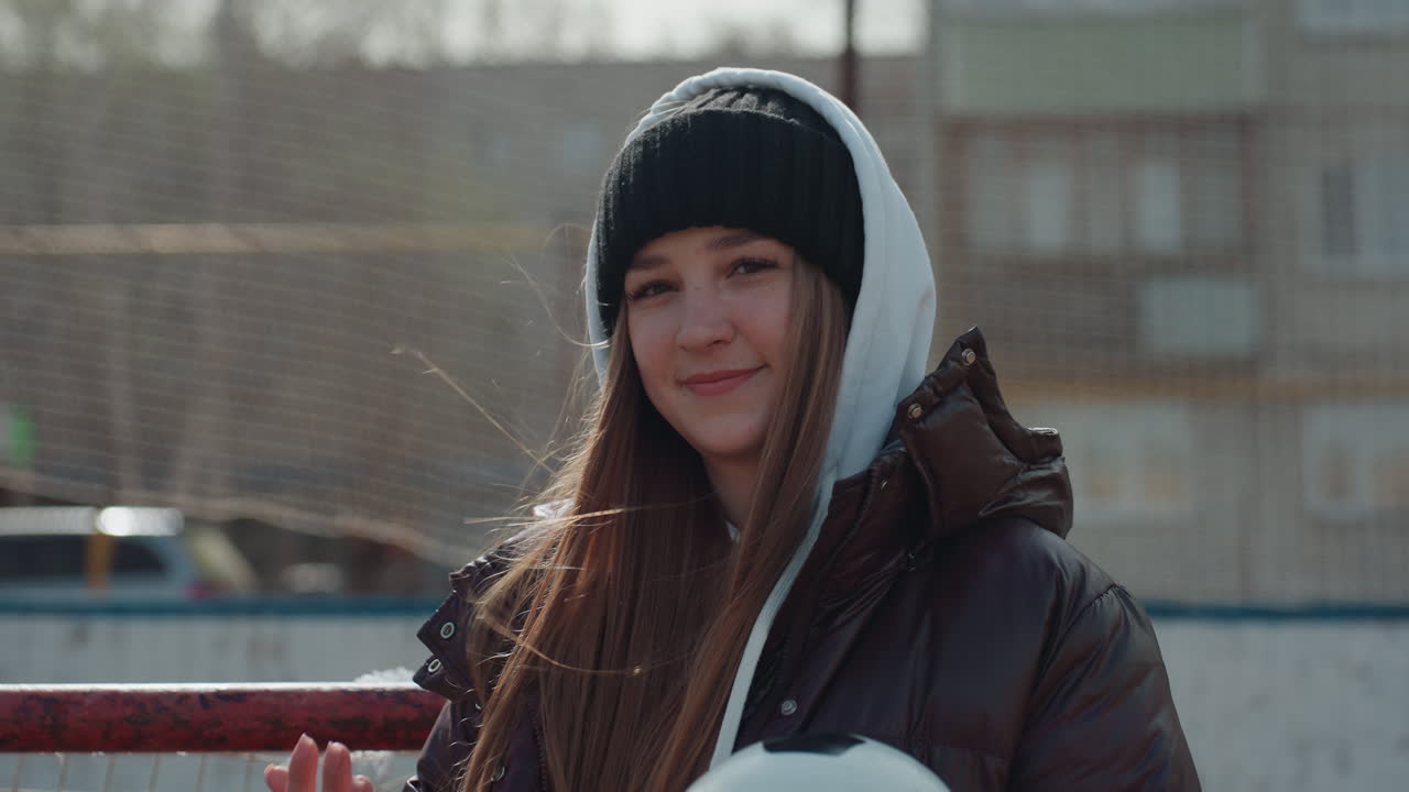 confident caucasian teen by soccer goal, hands steady on ball, wind-tossed hair and determined gaze, urban housing blocks behind net, poised stance suggests readiness for practice, cool daylight mood