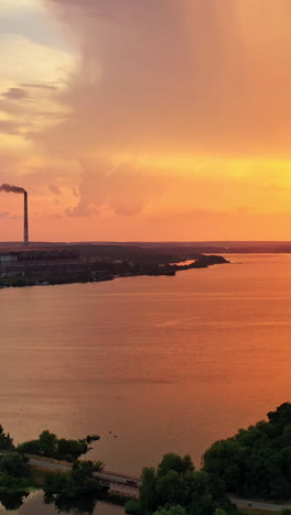Rural background near the lake at sunset. Aerial view of pipes with smoke at the bank of the river in the evening. Amazing sunset over the beautiful scenery. Vertical video