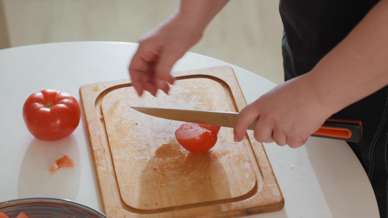Primo piano dall'alto di mani che affettano un pomodoro rosso maturo su un tagliere di legno in una cucina luminosa, mostrando un movimento stabile del coltello, una presa ferma della mano, succo fresco, semi e una preparazione pulita del cibo.