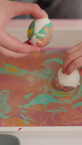 woman and little girl roll eggs in oily water with ebru decor mix in plastic tray closeup slow motion. easter holiday celebration ideas and traditions