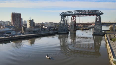 Aerial-pull-out-shot-reveals-a-boat-cruising-on-Riachuelo-river-with-Puente-Transbordador-Nicolas-Avellaneda-bridge-in-the-background,-in-La-Boca-neighbourhood