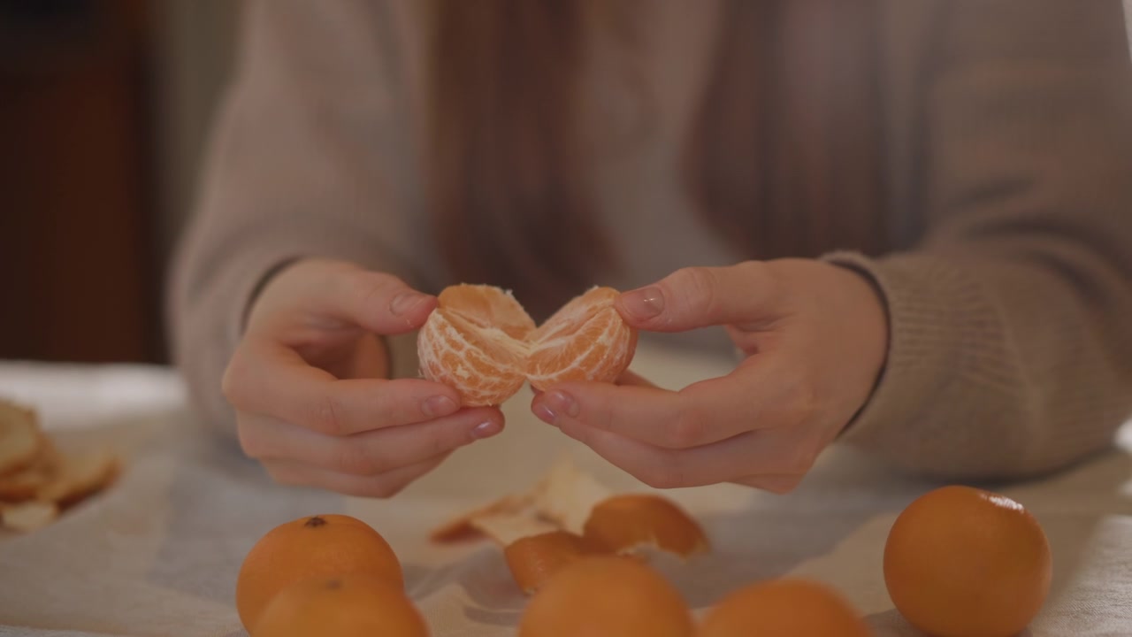 Tangan memegang belahan jeruk mandarin di atas meja, segmen berpasangan terpisah perlahan dengan jeruk mandarin utuh dan kulit di sekitarnya, manset sweter terlihat, pencahayaan lembut hangat, isyarat menawarkan dan rumah yang intim