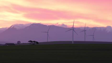 wind turbines slowly turning during sunset against a cloud capped mountain backdrop