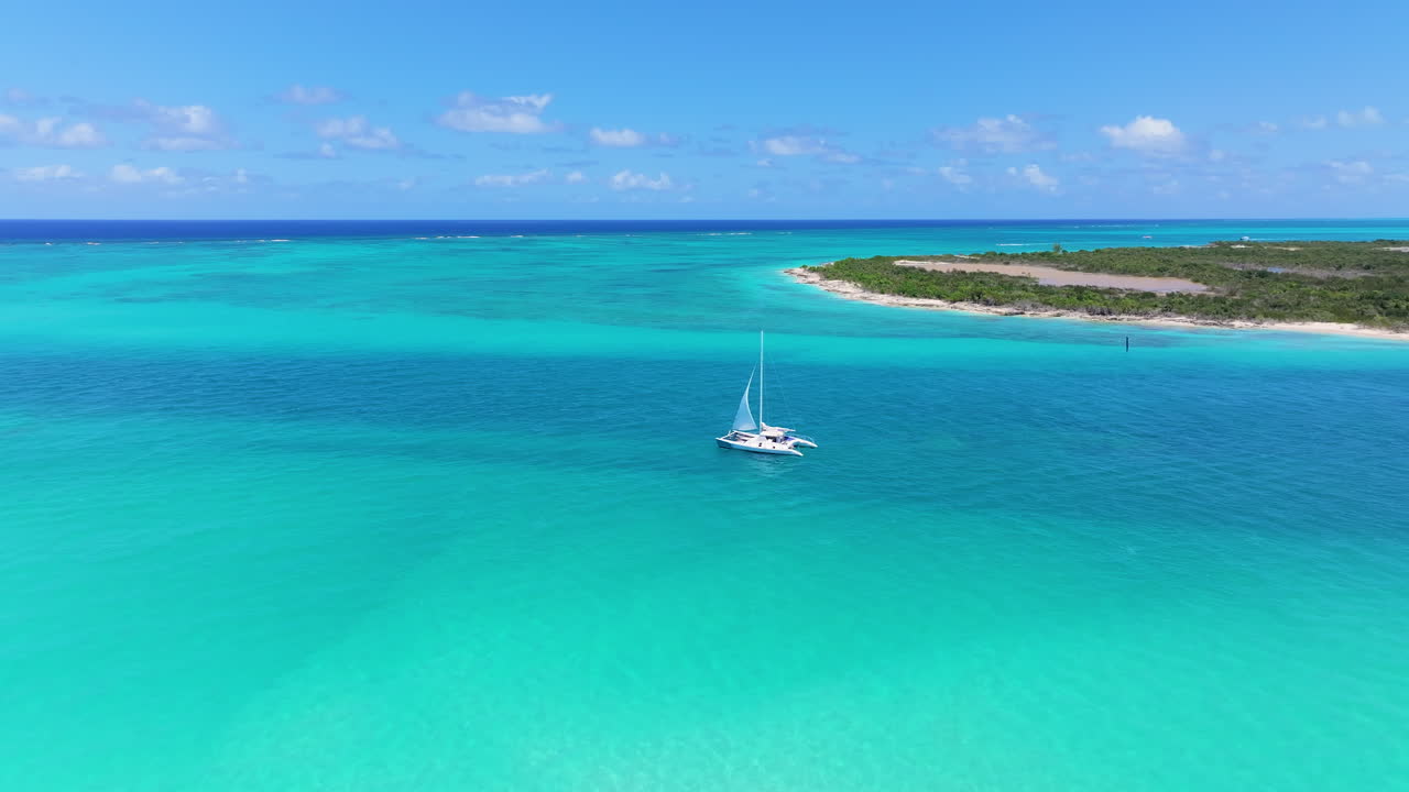 Serene forward shot of small sailboat gliding through the vast blue ocean. The calm waters extend towards the horizon, blending with blue sky with a small island with lush greenery on the right