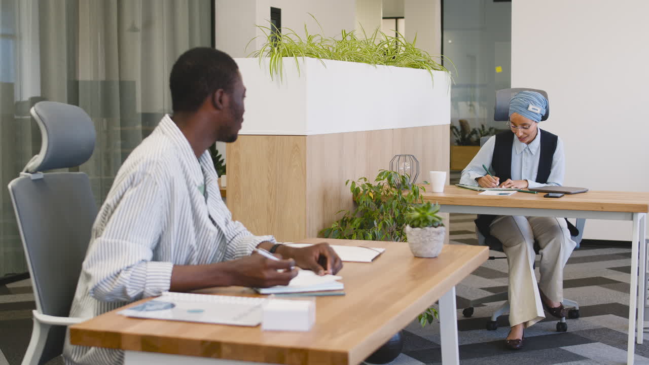Young Smiling Worker Working Taking Notes Sitting At His Desk And Talking With Muslim Businesswoman Who Works Sitting At Their Desk 1
