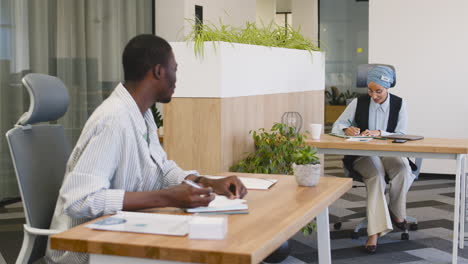 Young-Smiling-Worker-Working-Taking-Notes-Sitting-At-His-Desk-And-Talking-With-Muslim-Businesswoman-Who-Works-Sitting-At-Their-Desk-1