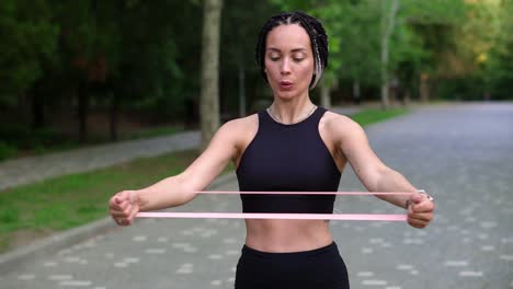 Young-athletic-girl-engaged-in-fitness-wearing-black-sportswear-with-a-rubber-band-in-her-hands-in-a-summer-park.-Girl-with