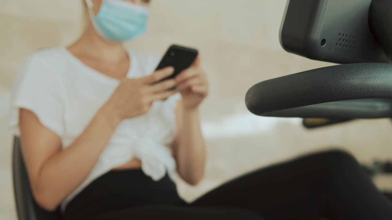 얼굴 마스크를 쓴 젊은 스포츠 여성은 체육관에서 스마트폰과 운동기를 사용합니다