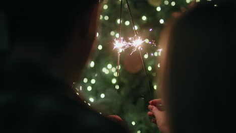 A-young-couple-holds-burning-sparklers-in-their-hands,-sits-facing-a-large-Christmas-tree-at-home