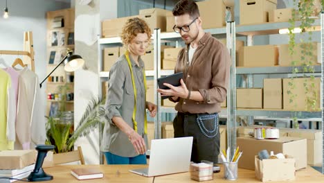 Caucasian-young-woman-designer-typing-on-laptop-and-writing-order-details-in-clothing-shop-warehouse-while-male-coworker-typing-on-tablet-and-checking-boxes