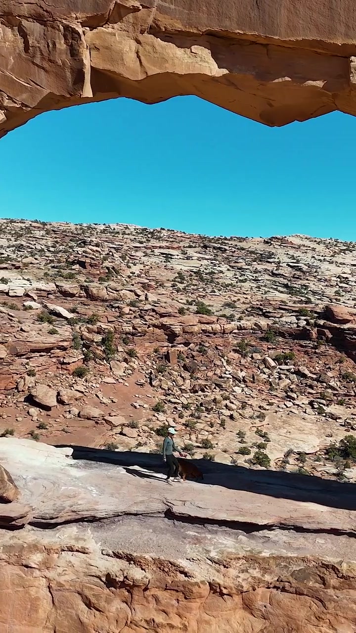 Ripresa verticale con drone di una donna e un cane che camminano sotto il suggestivo Jeep Arch, Moab, Utah, USA