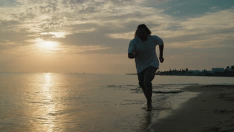 Plano-Medio-De-Un-Joven-Corriendo-En-La-Playa-En-El-Agua-Al-Amanecer-En-Cámara-Lenta