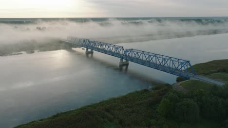 aerial establishing view of the steel bridge over lielupe river on a sunny summer morning, fog rising over the river, cars driving, wide drone shot moving forward, tilt down