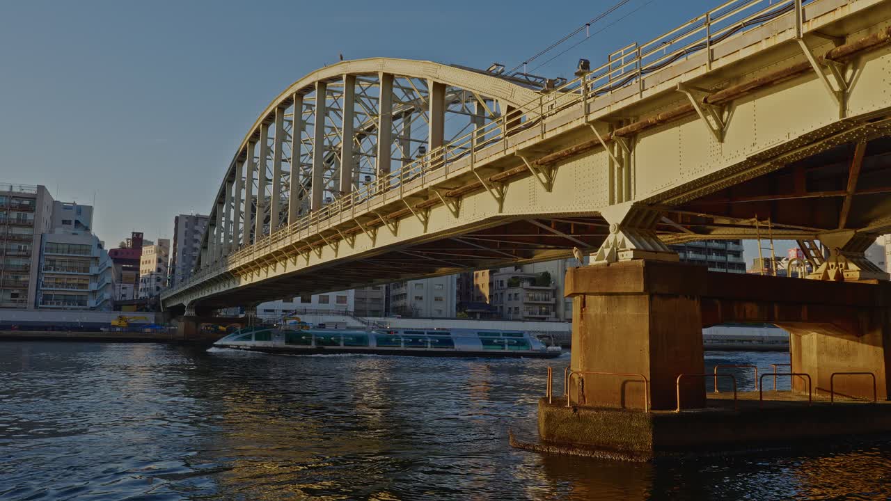 A wide shot of the JR Sobu Line Sumidagawa Railway Bridge, a historic steel arch bridge in Tokyo, with a water bus passing underneath on the Sumida River at sunset