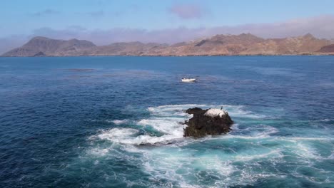 a rocky outcrop near cedros island surrounded by foamy waves and deep turquoise waters, drone approach with boat floating in distance
