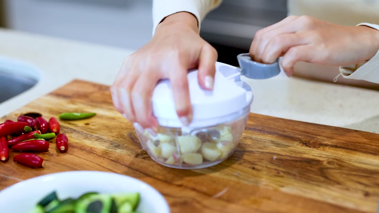 Hands using a manual food chopper to prepare garlic on a wooden board in a bright kitchen setting
