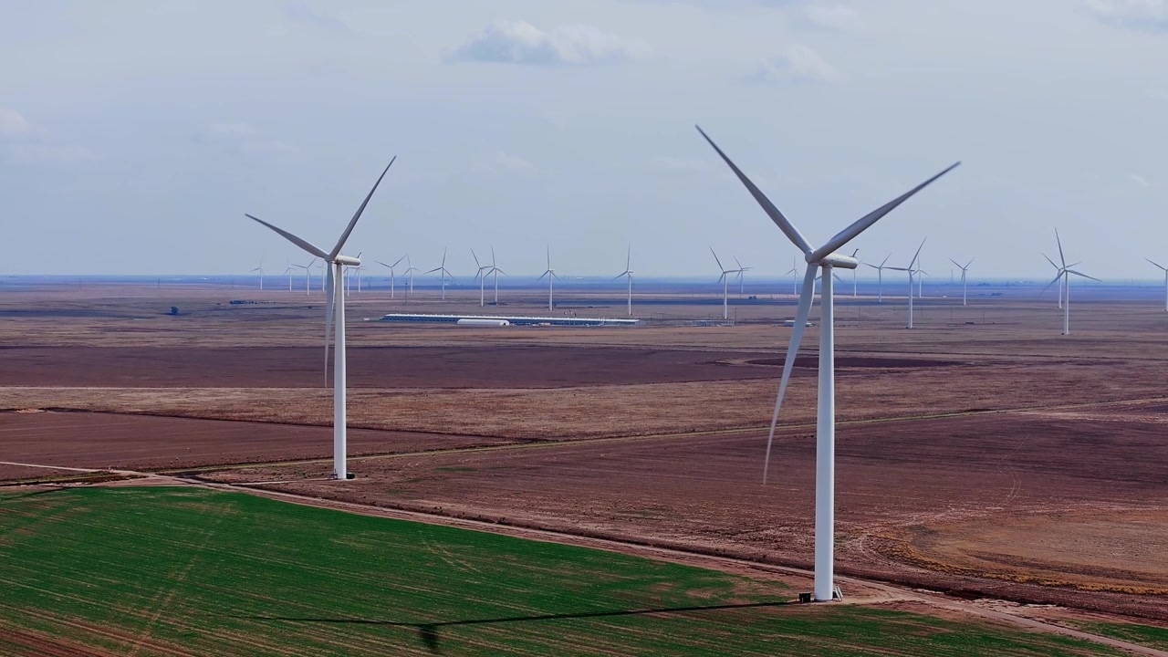 Vista aérea estática de 2 grandes turbinas eólicas giratórias acima de terras agrícolas do Texas em tons de verde e marrom, como um mosaico, com um vasto parque eólico comprimido em direção ao horizonte sob céus nebulosos