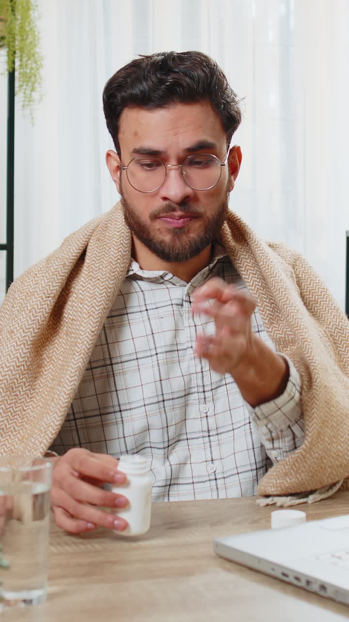 un hombre de negocios indio enfermo envuelto en una manta trabaja en una computadora portátil en el escritorio de la oficina de casa toma medicamentos
