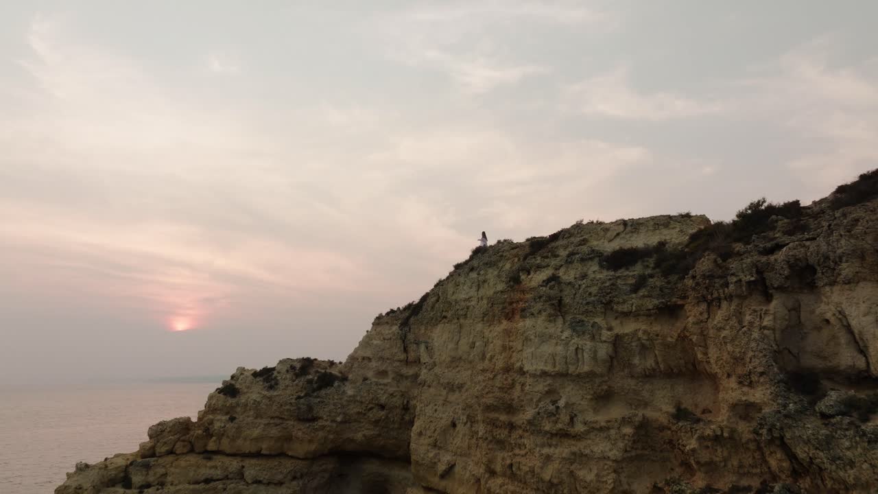 A woman gazes at the hazy sunset from a rocky cliff, surrounded by soft skies and calm ocean views in Albufeira, Portugal