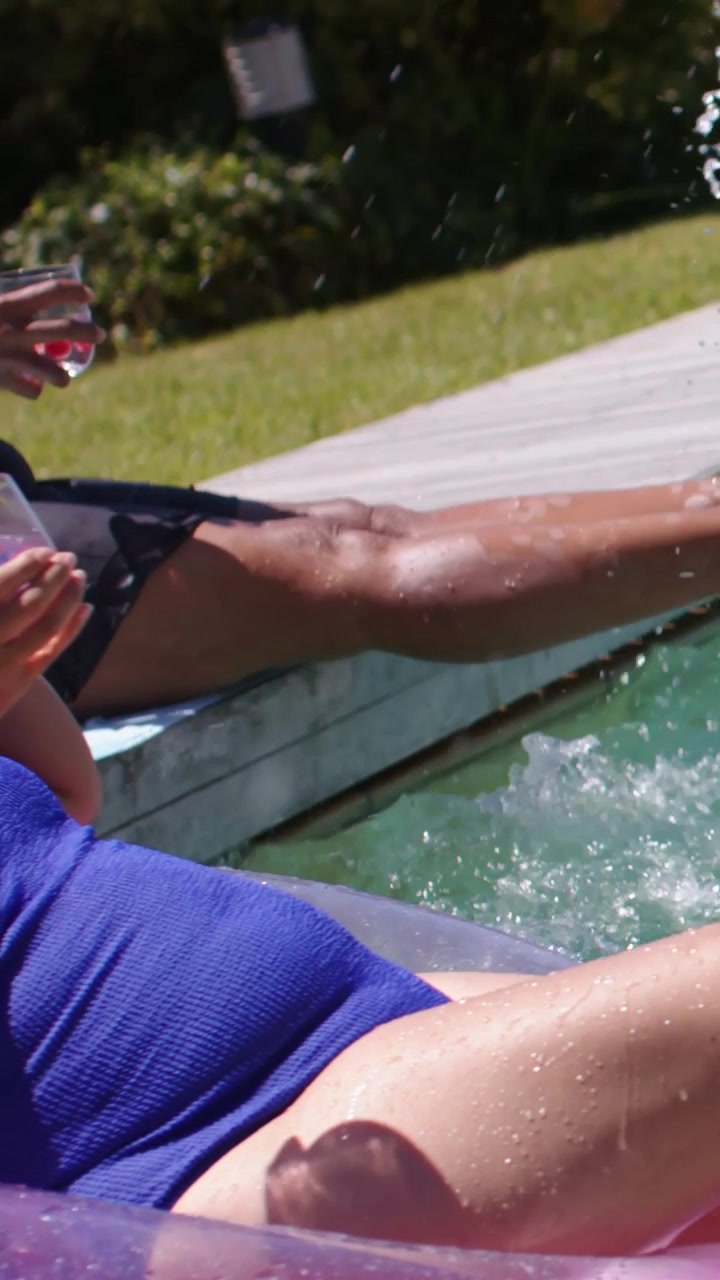 Vertical video: Sun urging senior in swimsuit kicking at pool edge, splashing woman with pink drink