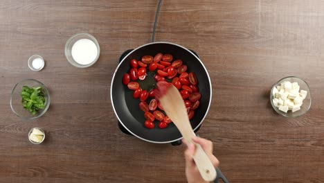 Top-Down-Schuss-Von-Kirschtomaten,-Die-In-Einer-Pfanne-Auf-Einem-Tischkochfeld-Auf-Einem-Holztisch-Mit-Anderen-Zutaten-Sautiert-Werden,-Tomatenmark-Hinzufügen-Und-Umrühren