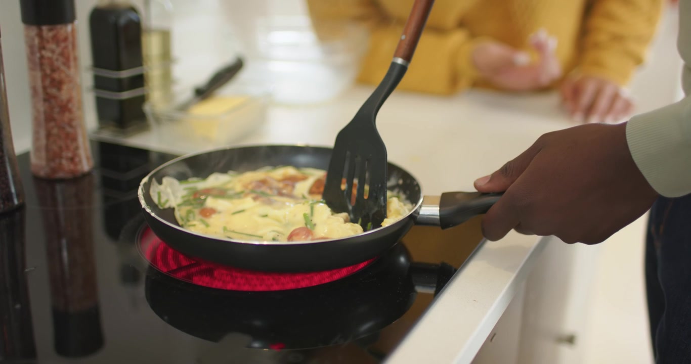 Un couple observant une poêle posée sur une plaque de cuisson chaude pendant qu'un homme cuisine une omelette, pliant les œufs avec une spatule.