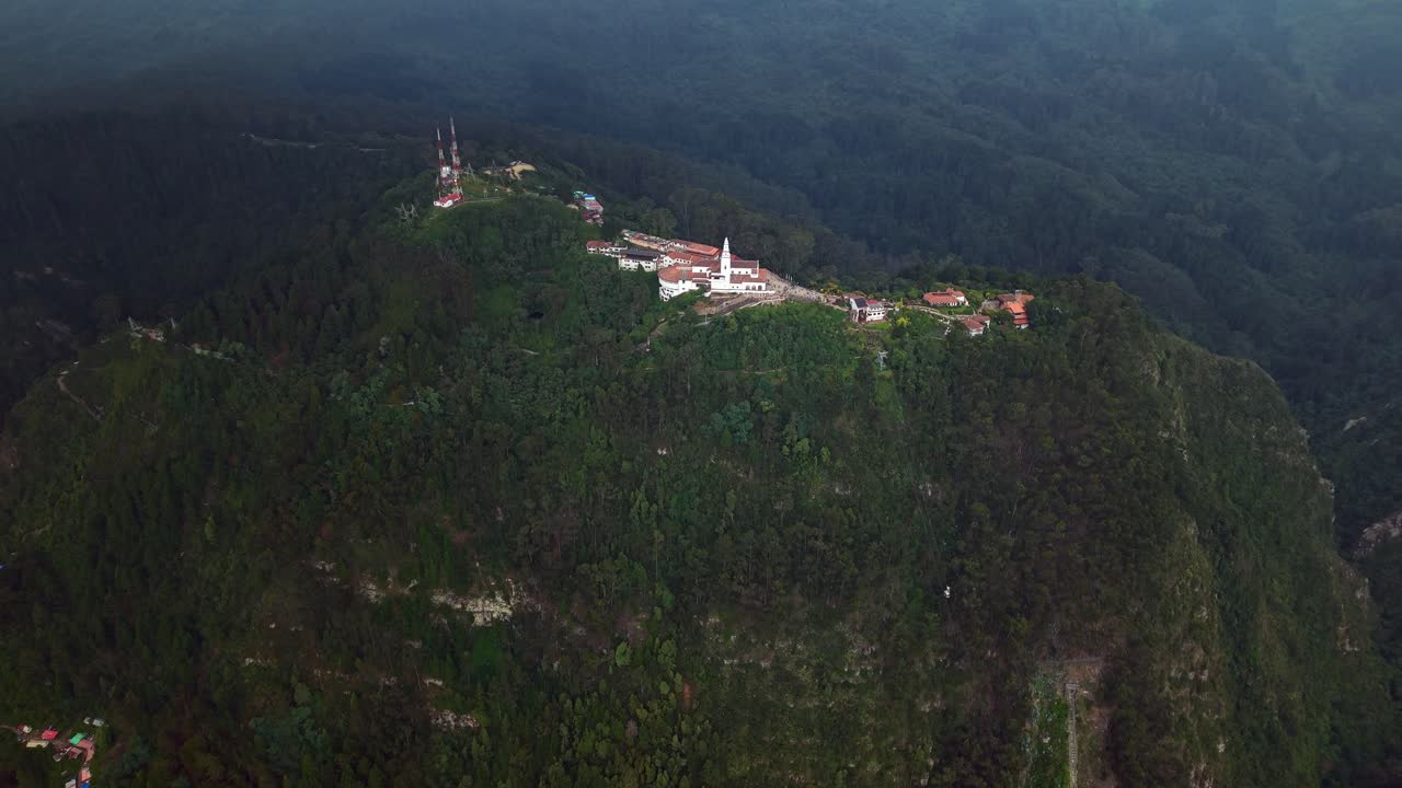 Aerial drone approach to the iconic Monserrate Sanctuary in Bogotá, Colombia, flying above the church and surrounding buildings on a hilltop covered in dense green trees and vegetation