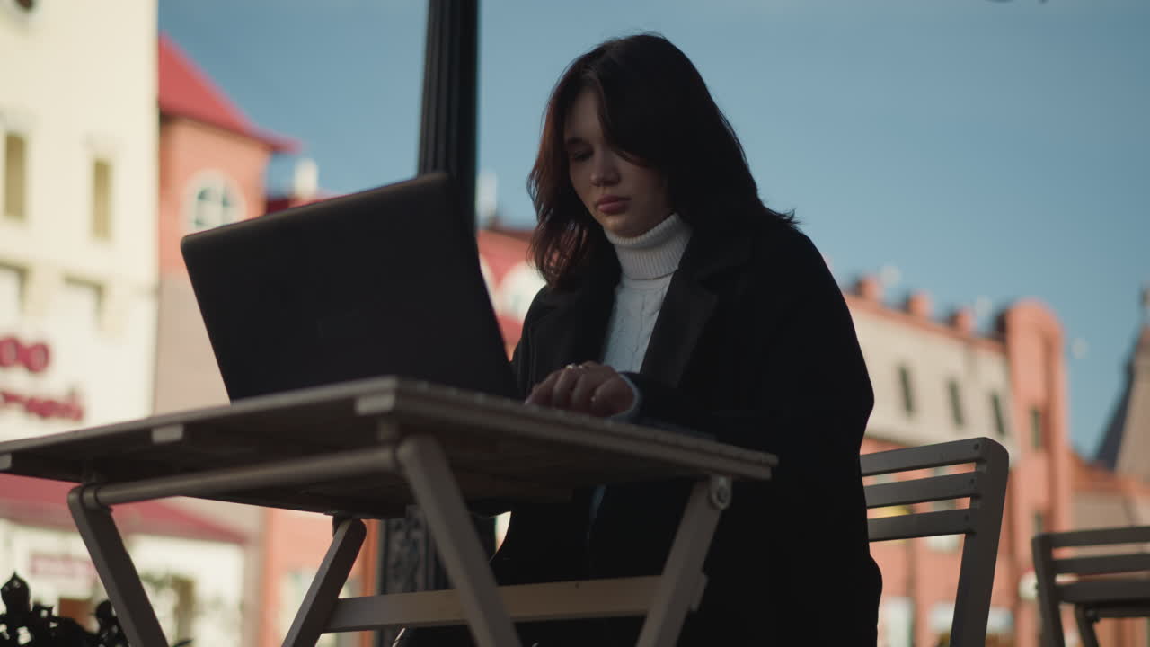 Lady working on laptop outdoors in urban setting, wearing stylish black coat , with decorative black pillar nearby, blurred red brick building in background, and flower swaying gently in the breeze
