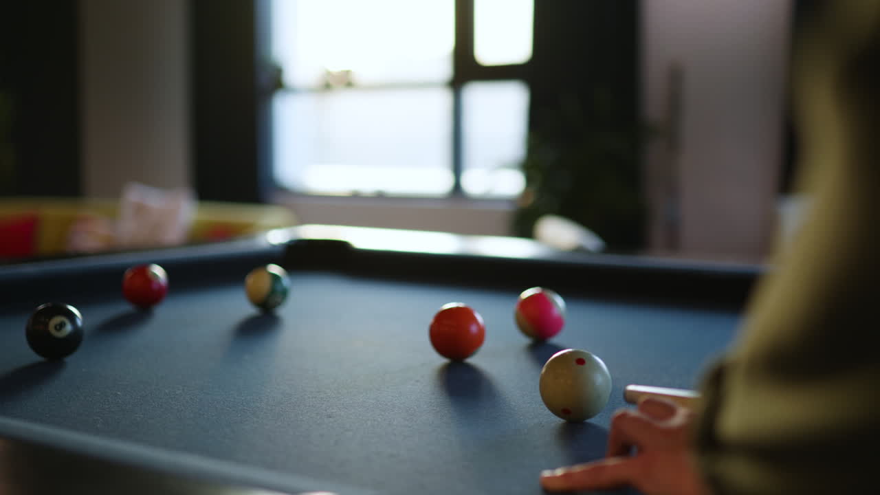 A person enjoys a game of billiards in a modern lounge. Colorful balls are arranged on a pool table, illuminated by warm afternoon light. Comfortable seating surrounds the area.