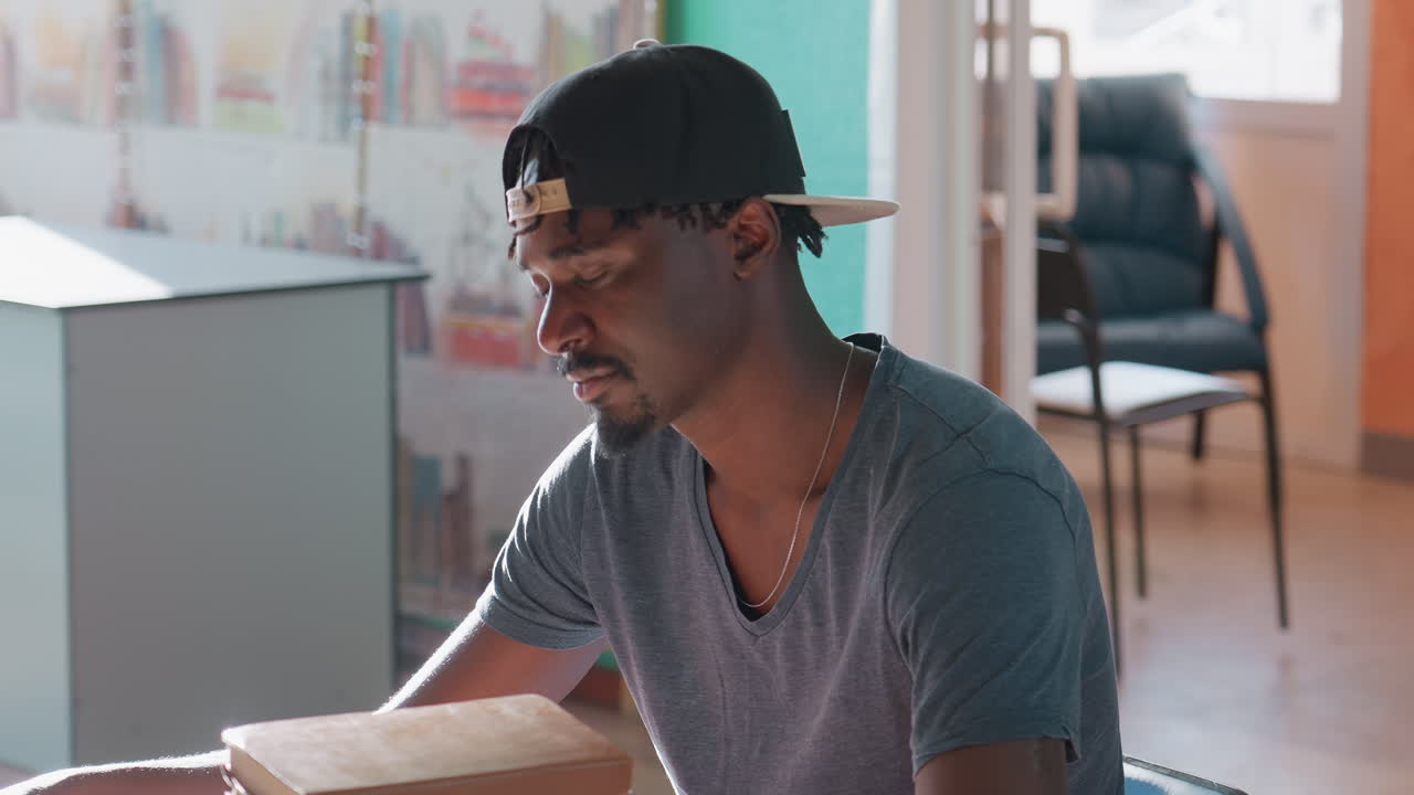 Man in cap sits at library table beside tall stack of books, arms resting, notebook and pen nearby, warm sunlight filling modern study room with shelves, chairs, and colorful wall in background