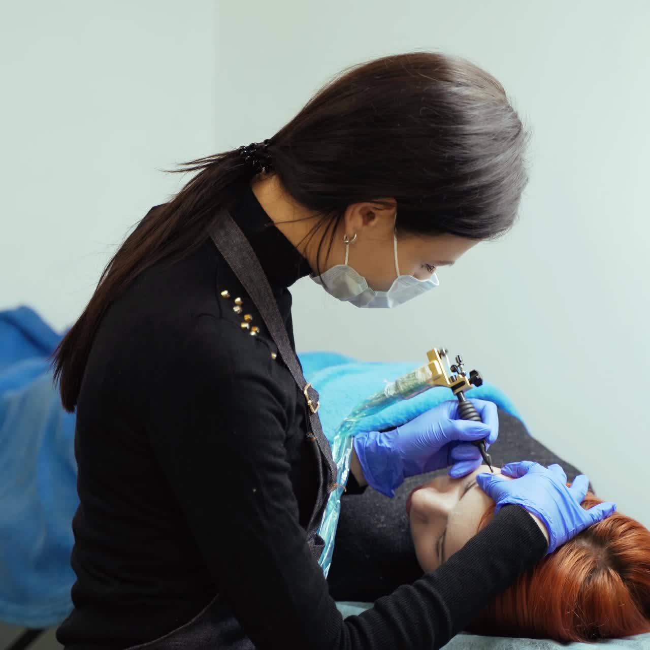 A young woman in gloves and a mask is performing eyebrow tattoo for client who laying on the couch in a beauty salon. Permanent makeup. Beauty industry