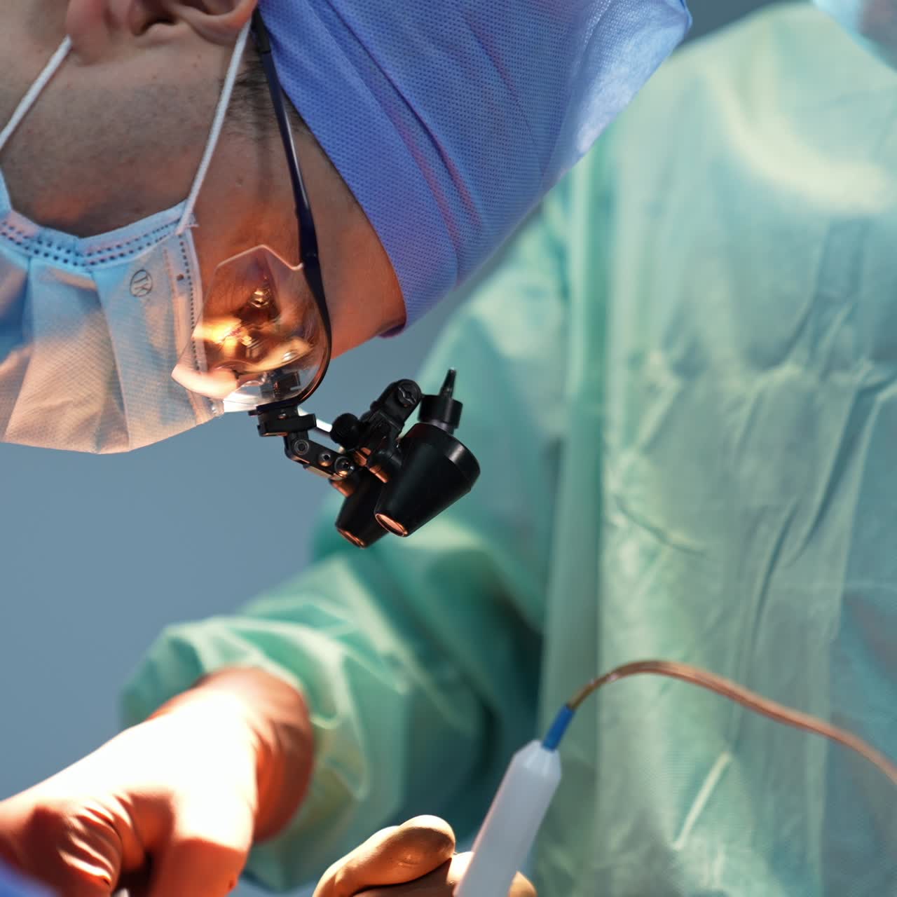 Chief surgeon in mask, cap and device glasses bent his head low over the patient. Close up portrait of a specialist focused on surgery