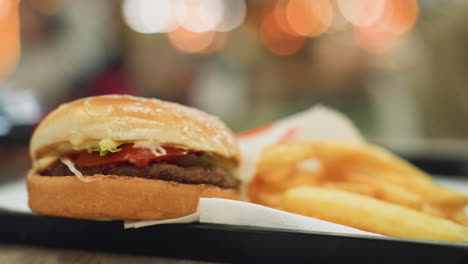 patatas fritas doradas y una hamburguesa en una bandeja negra en una mesa con un fondo borroso de luces, enfatizando la deliciosa comida rápida en un entorno de comedor vibrante