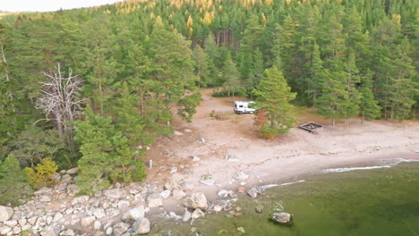 Sweden---White-Campervan-Nestled-in-Swedish-Nature,-Surrounded-by-Autumn-colored-Fir-and-Birch-Trees,-With-the-Sea-Stirred-by-the-Wind-on-an-October-Morning---Aerial-Pan-Right