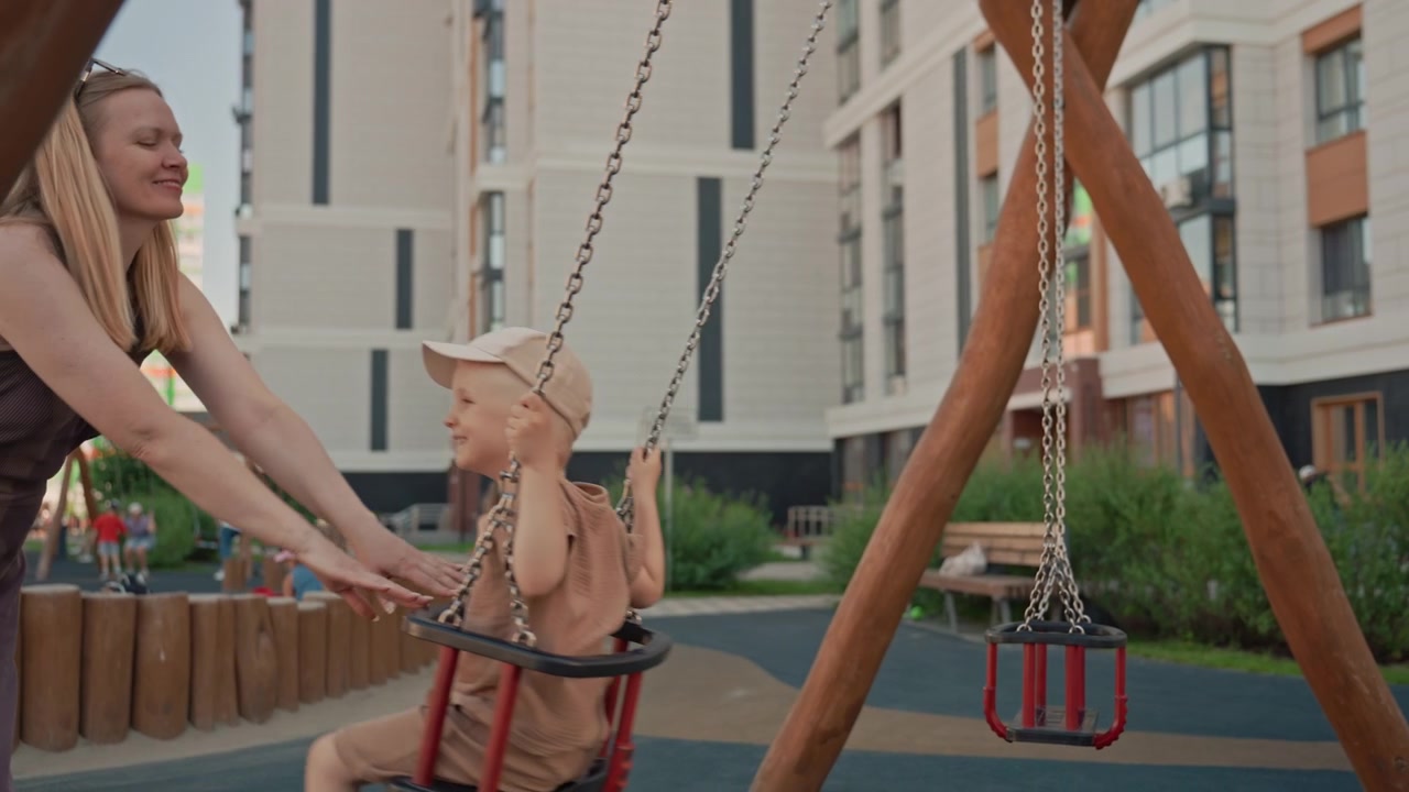 Una donna spinge delicatamente un bambino sull'altalena in un parco giochi all'aperto. La scena cattura l'interazione tra loro, con altri bambini che giocano sullo sfondo vicino a strutture di gioco in legno e moderne.